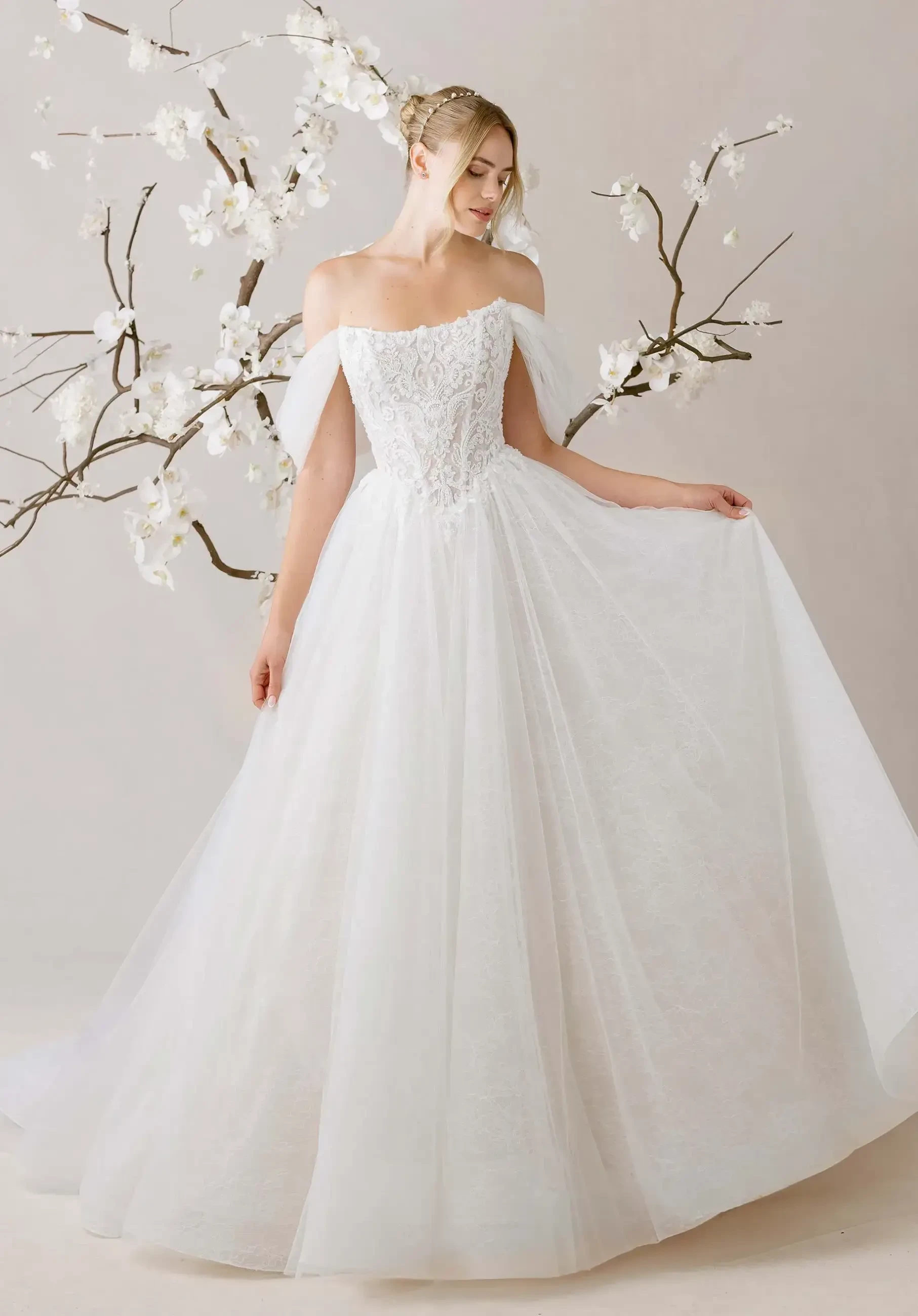 A woman in an off-shoulder white wedding gown stands gracefully, holding the skirt. Orchids extend behind her, creating a serene, elegant scene.