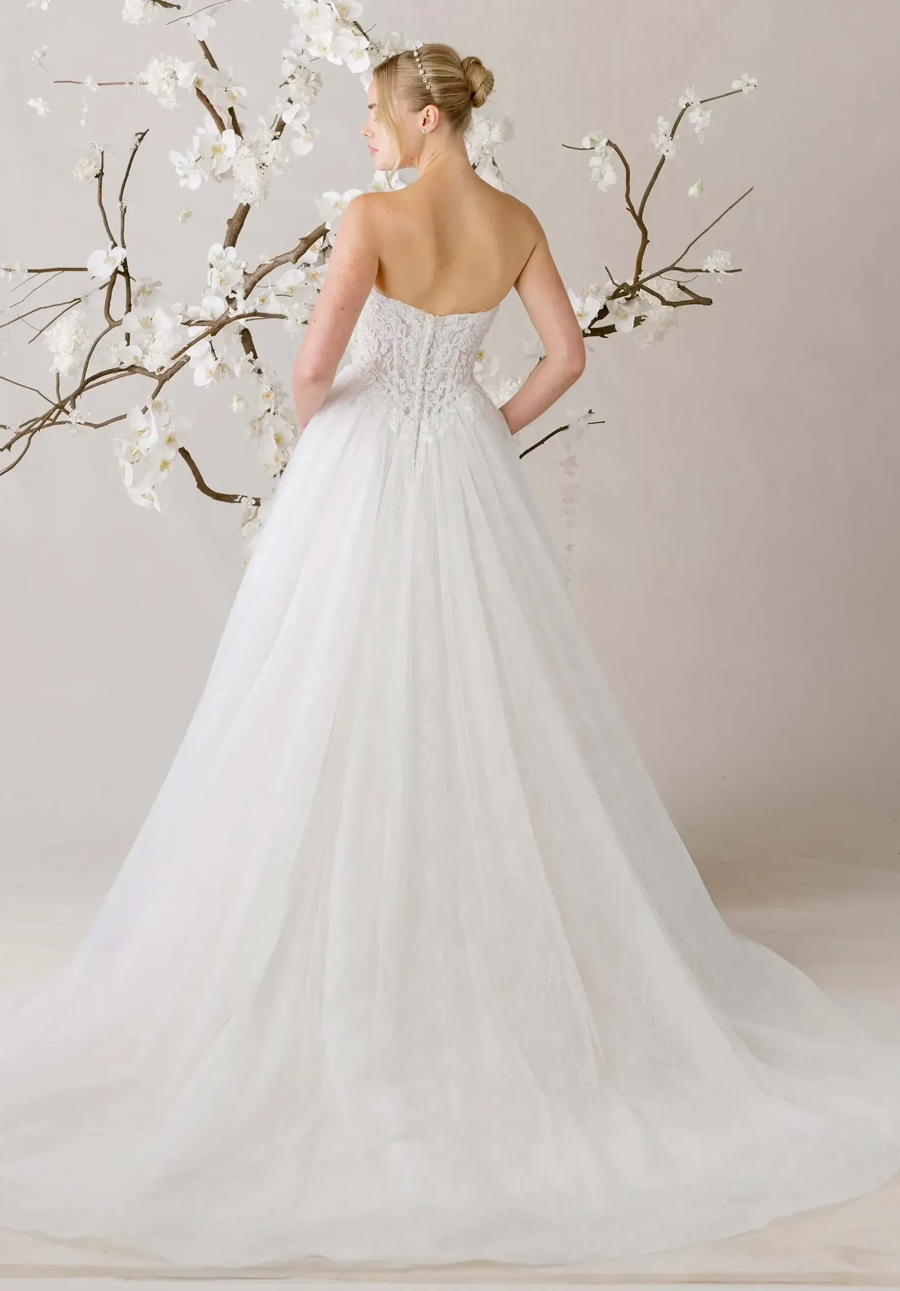 A bride in a strapless white gown stands with her back to the camera, near delicate, blooming branches. The scene conveys elegance and serenity.