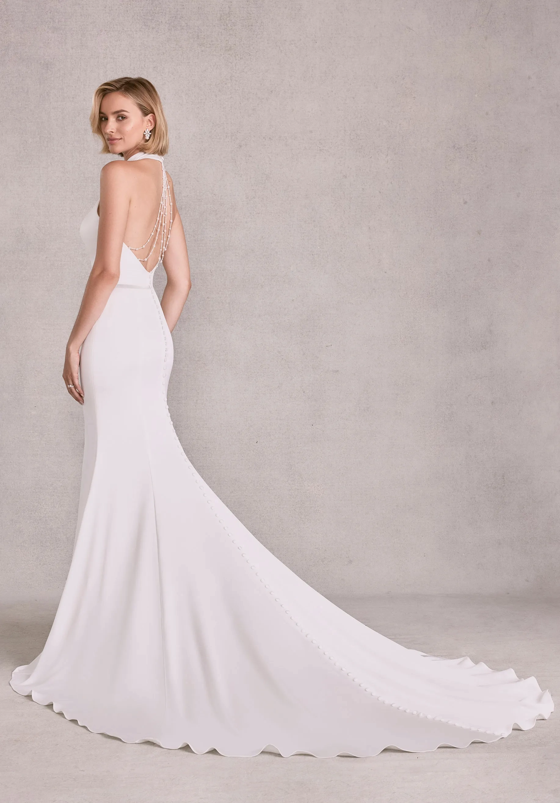 Model is wearing a fit and flare wedding gown with a collar style halter neckline and open back with crystal and pearl chandelier beading in crepe back satin