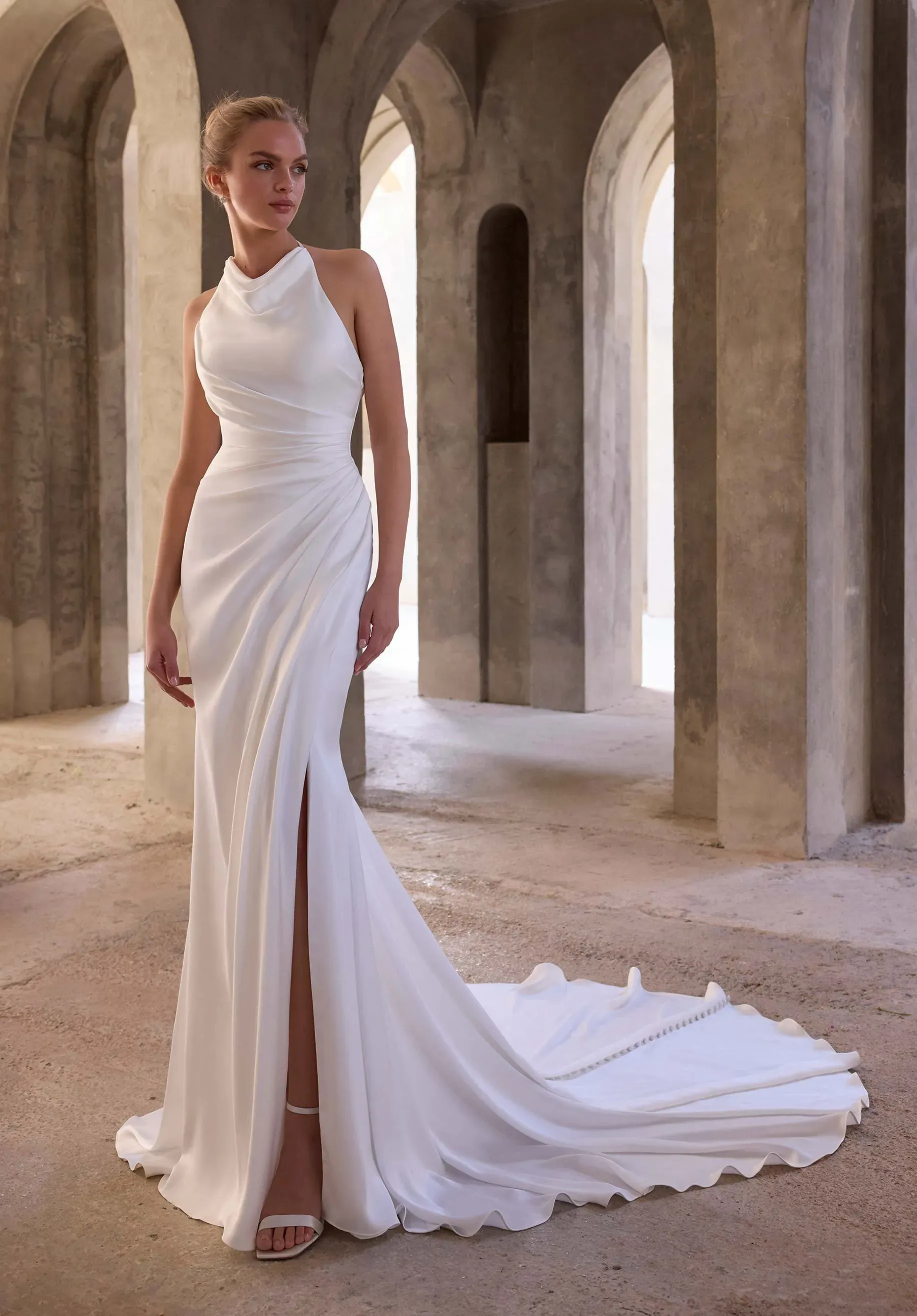 Model is wearing a high neck halter sheath wedding dress with a satin skirt and slit