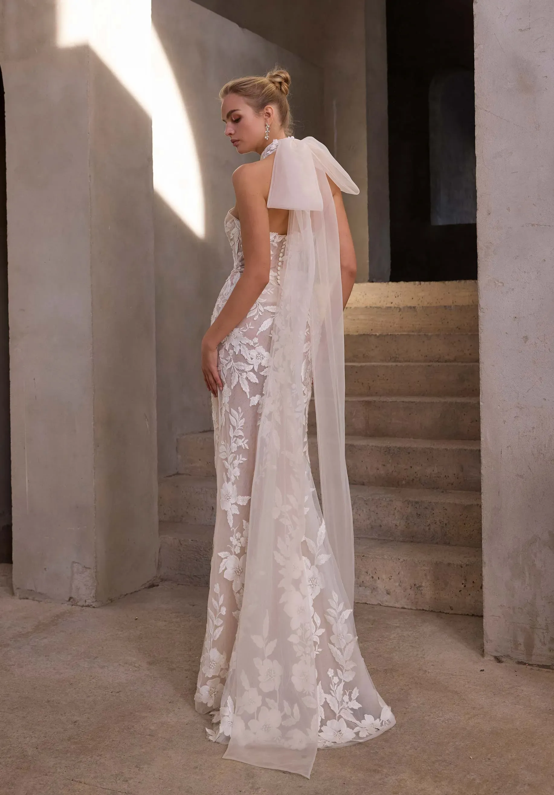 Model is wearing a sheath bridal gown with a halter neckline made of floral appliqued tulle and a detachable organza bow with blouson sleeves