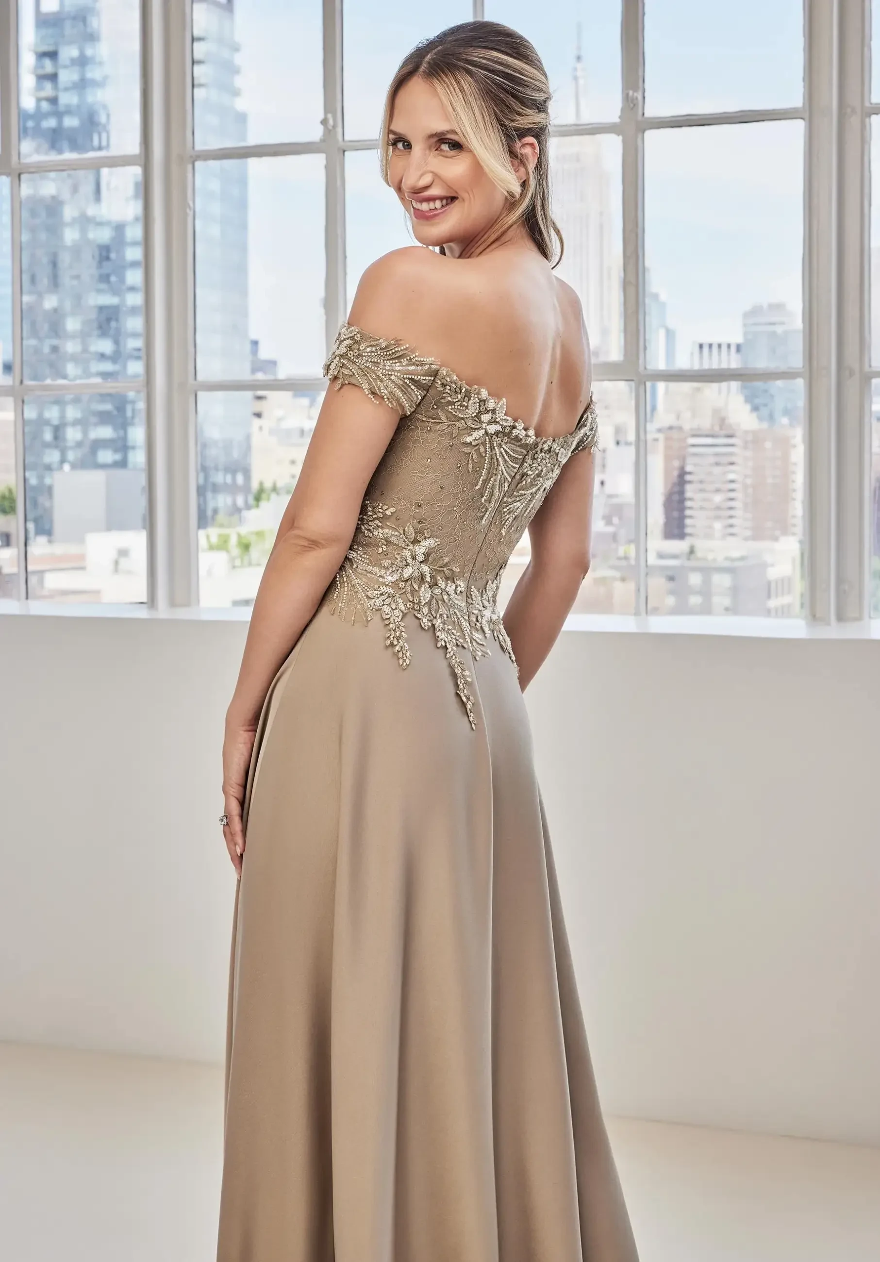 Model is wearing an a-line evening dress with an off the shoulder neckline and botanical embroidered bodice in crepe