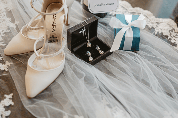 Elegant bridal accessories display: ivory heels, sparkling necklace and earrings in a black box, teal gift box with white ribbon on lace fabric.