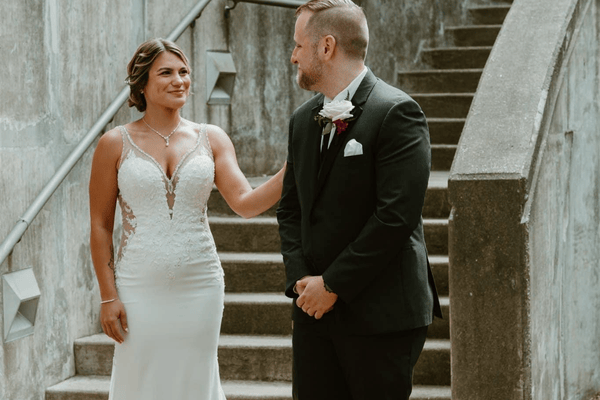 A bride in a lace gown and a groom in a black suit share a tender moment on stone steps. Both are smiling warmly, conveying joy and affection.