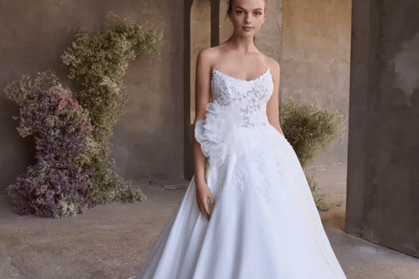 A woman in an elegant white wedding gown adorned with lace and a large floral detail stands in a rustic setting, surrounded by soft pastel flowers.