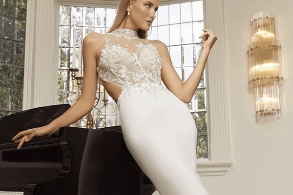 Elegant woman in a form-fitting white gown with intricate lace details leans against a grand piano. Softly lit room with a large window and modern wall light.