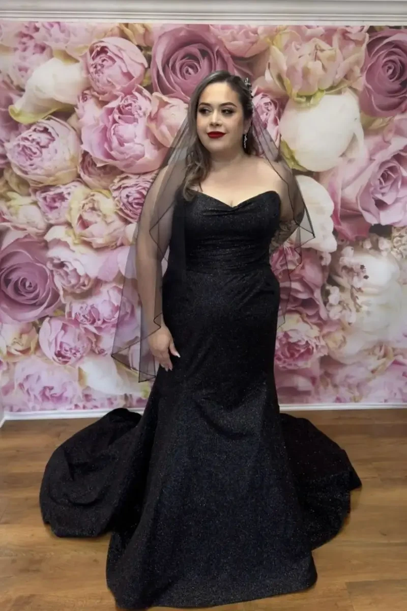 Bride wearing a black strapless wedding gown with a fitted silhouette and long train, standing in front of a pink floral backdrop with a sheer black veil.
