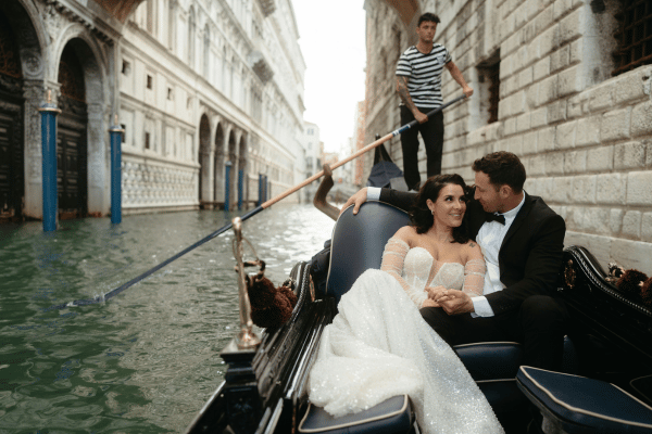 Bride and groom in a destination wedding gown and tuxedo riding a gondola through a Venice canal.