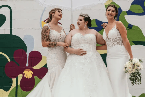 Three brides in lace wedding dresses posing together outdoors against a colorful mural