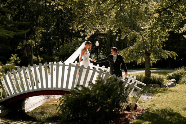 Bride and groom crossing a garden bridge, groom holding the bride&rsquo;s hand, surrounded by greenery at the Friendship Botanic Gardens