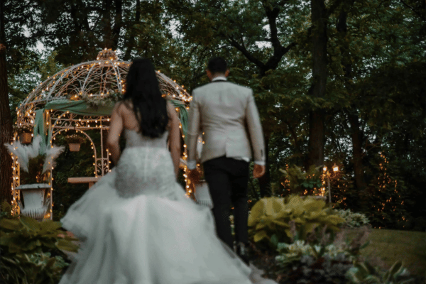 Back view of the bride and groom walking toward a gazebo with string lights, trees, grass, and plants