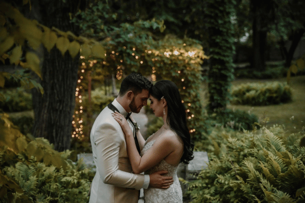 Bride and groom standing with their heads leaning together in a garden with greenery and string lights, groom&rsquo;s hands on the bride&rsquo;s waist and bride&rsquo;s hands on the groom&rsquo;s chest
