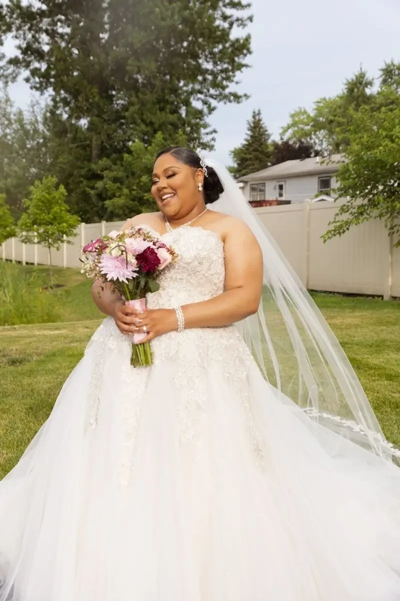 Bride in an embellished lace ball gown holding a pink and burgundy bouquet, smiling outdoors with her long veil flowing behind her.