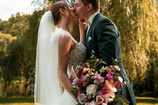The bride wearing her wedding gown and veil while holding her bouquet kissing the groom