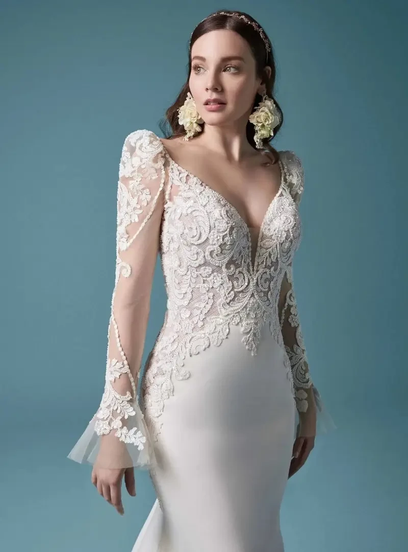 Model wearing a fitted wedding dress with a lace V-neck bodice, illusion long sleeves, and delicate floral detailing.