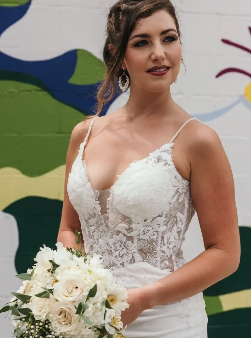 Bride wearing a fitted wedding dress with a floral lace bodice, plunging neckline, spaghetti straps, and white floral bouquet.