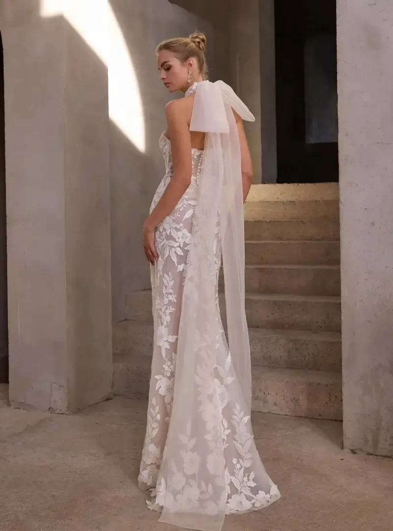 Back view of a fitted floral lace wedding dress with an open back, covered buttons, and a long sheer bow detail.