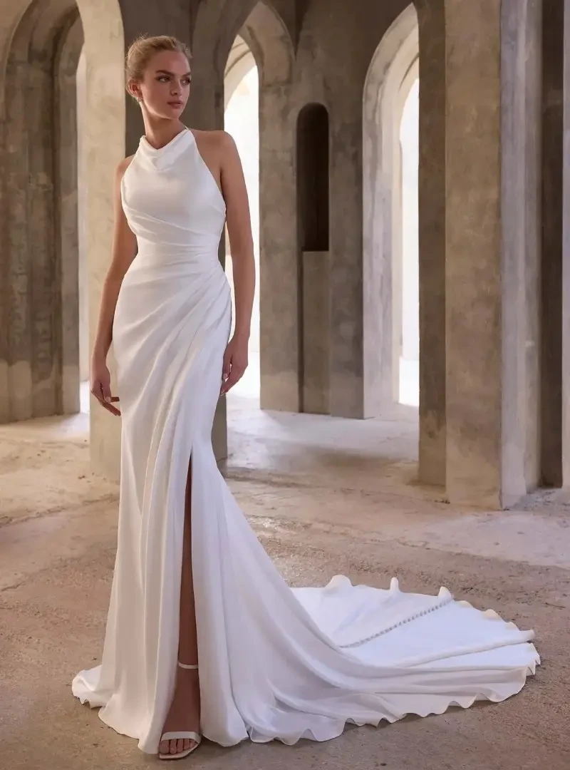 Model wearing a sleek fitted wedding dress with a halter neckline, ruched waist, front slit, and flowing train.